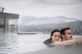 Your hotels in South Tyrol: the home of abundance Couple relaxing in outdoor pool with mountain view on a cloudy day.