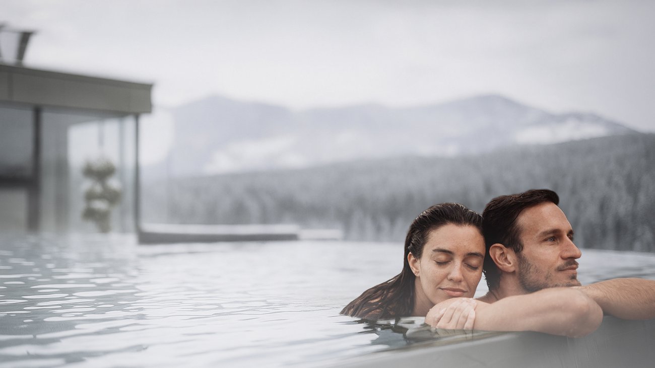 Your hotels in South Tyrol: the home of abundance Couple relaxing in outdoor pool with mountain view on a cloudy day.