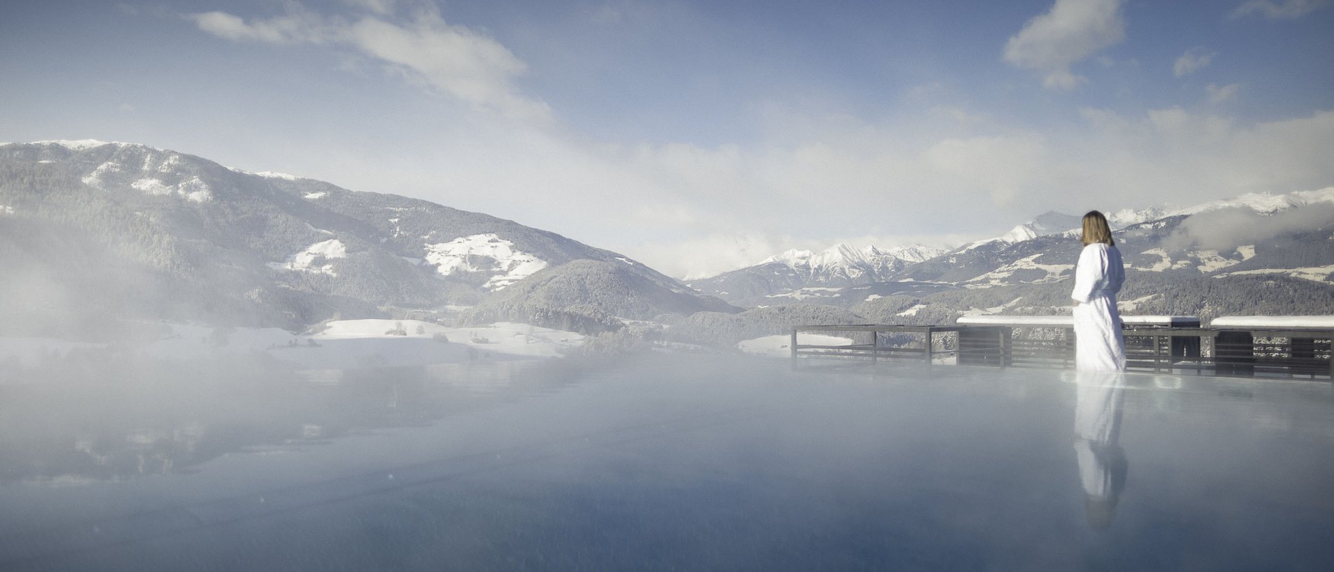 Die Quelle der Erfüllung: Ihr Hotel am Kronplatz Person in Bademantel steht am dampfenden Pool mit Bergblick im Winter