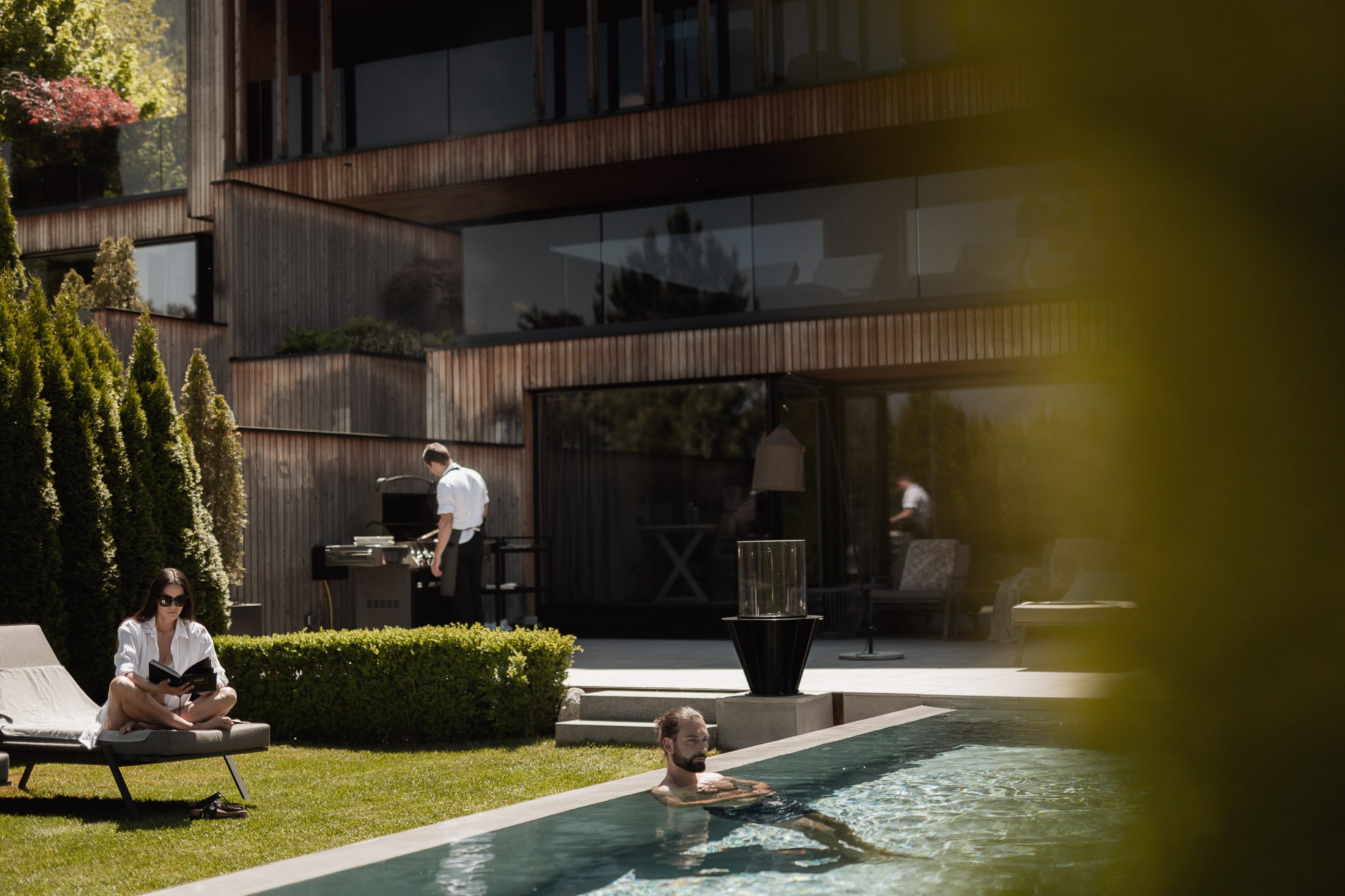 Erfüllender Urlaub in Ihrem Luxus-Chalet in Südtirol Person liest auf Liegestuhl, Mann schwimmt im Pool, Koch am Grill vor modernem Haus