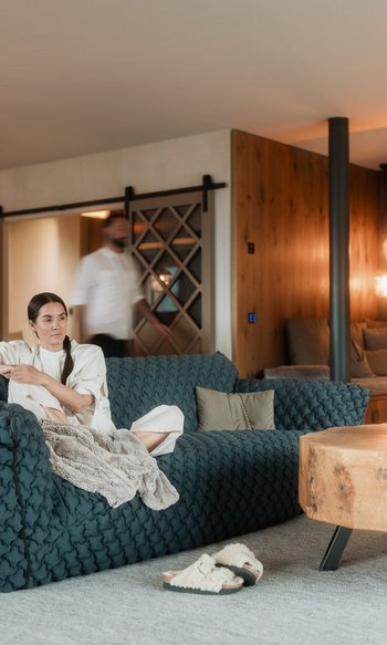 Your hotels in South Tyrol: the home of abundance Woman sitting on blue sofa in modern living room with wooden table and fireplace
