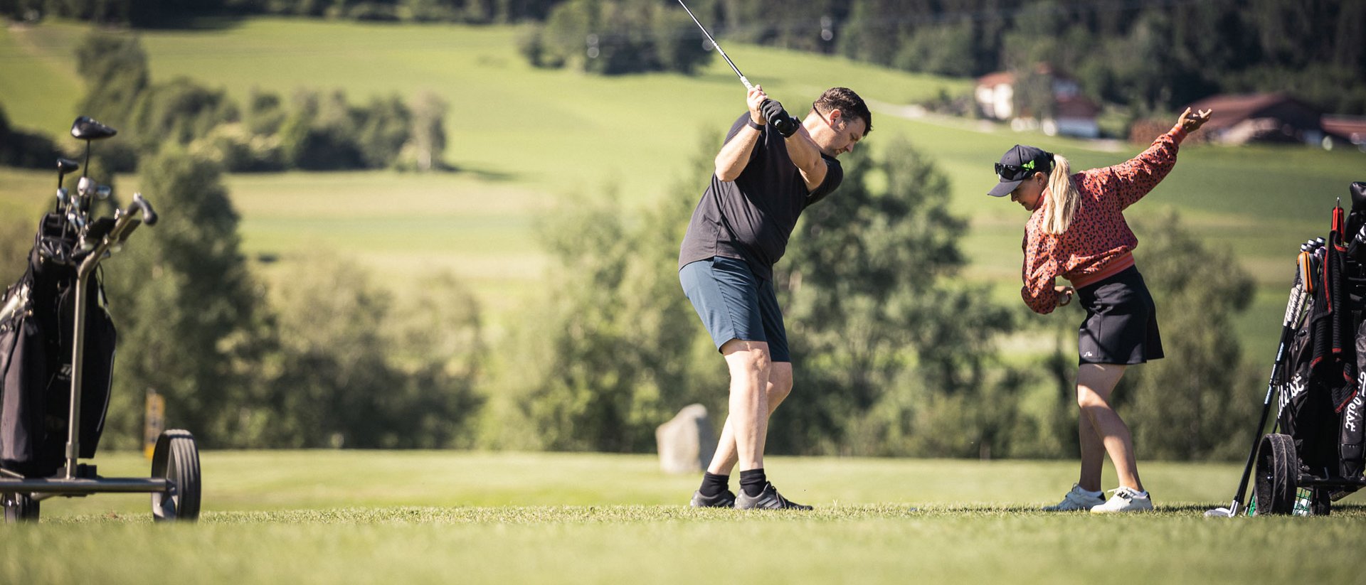 Die Quelle der Erfüllung: Ihr Hotel am Kronplatz Mann beim Golfabschlag mit Golfschläger, Frau zeigt Golfschwung auf grünem Feld