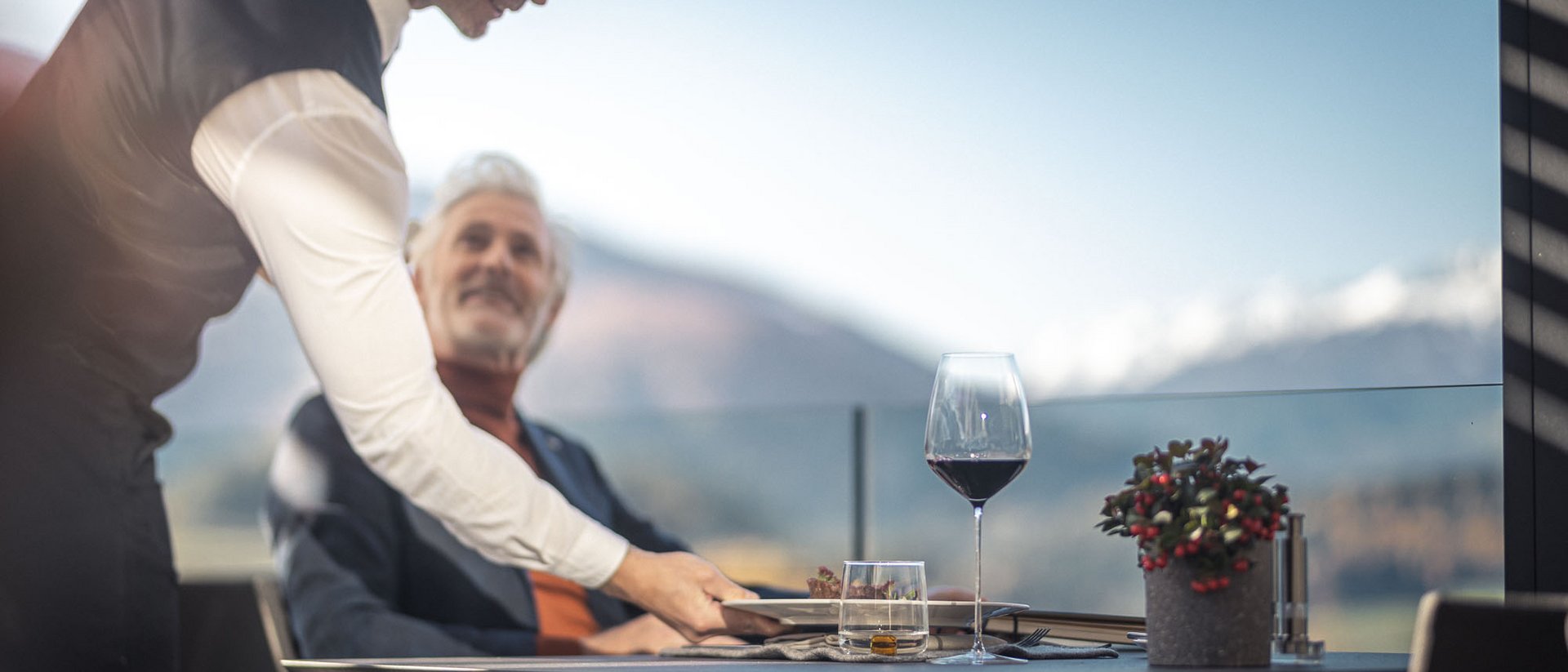 Die Quelle der Erfüllung: Ihr Hotel am Kronplatz Kellner serviert Essen an Tisch mit Wein und Blumen auf Terrasse bei Tageslicht