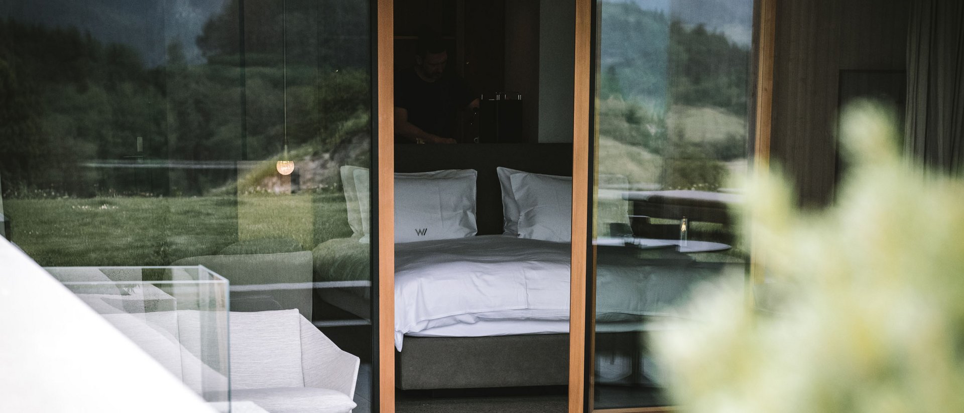 Die Quelle der Erfüllung: Ihr Hotel am Kronplatz Blick durch Glas auf ein modernes Hotelzimmer mit Bett und Berglandschaft