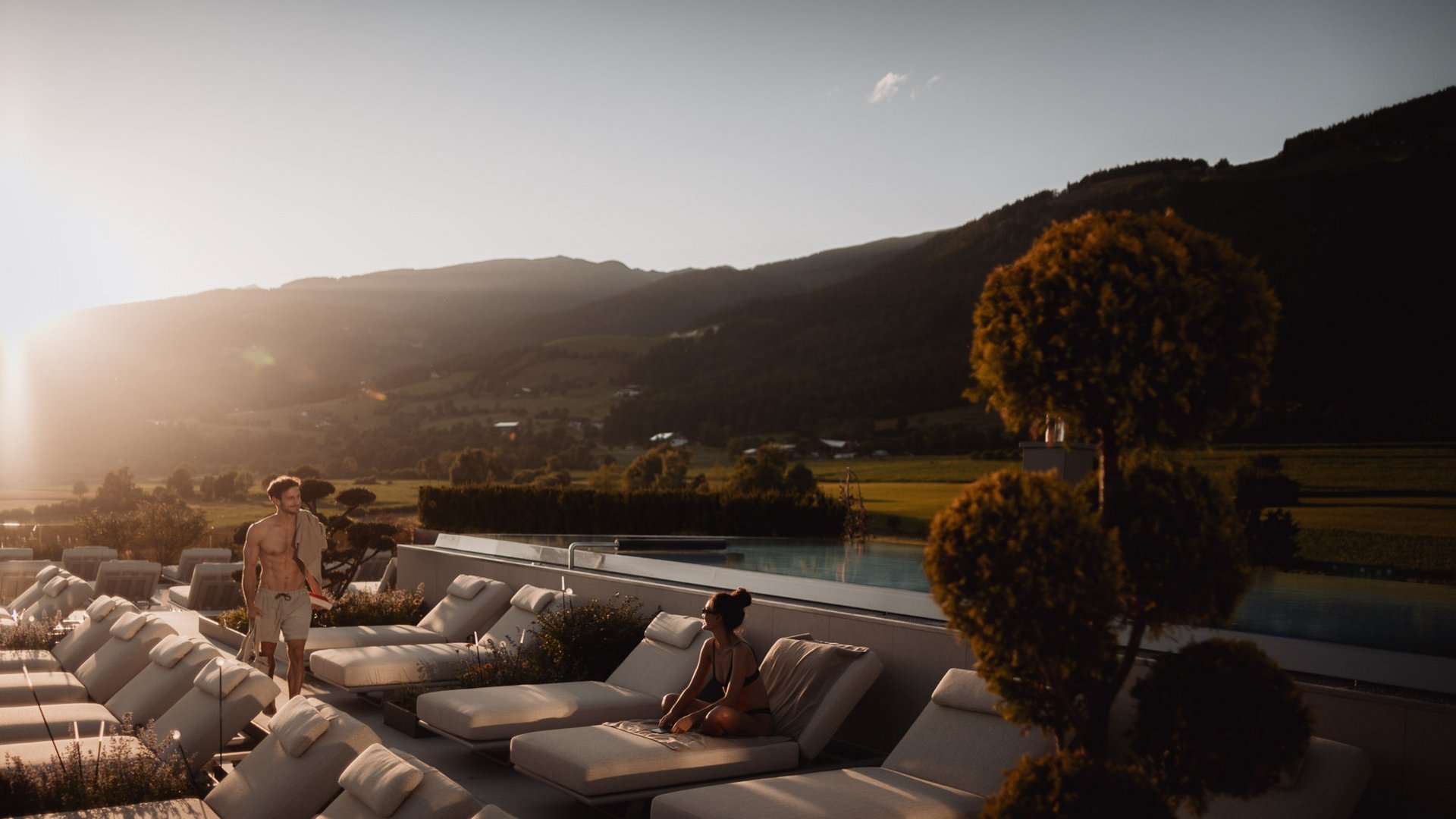 Your hotels in South Tyrol: the home of abundance People relaxing on lounge chairs by pool in sunny mountain landscape