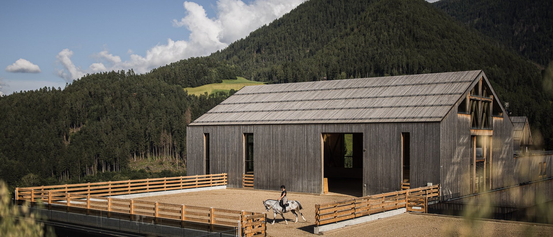 Erfüllender Urlaub in Ihrem Luxus-Chalet in Südtirol Reiter auf Pferd vor modernem Holzstall in Berglandschaft