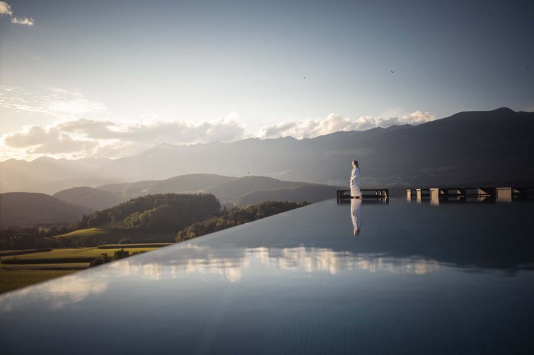 Die Quelle der Erfüllung: Ihr Hotel am Kronplatz Frau in Bademantel am Rand eines Pools mit Berglandschaft im Hintergrund