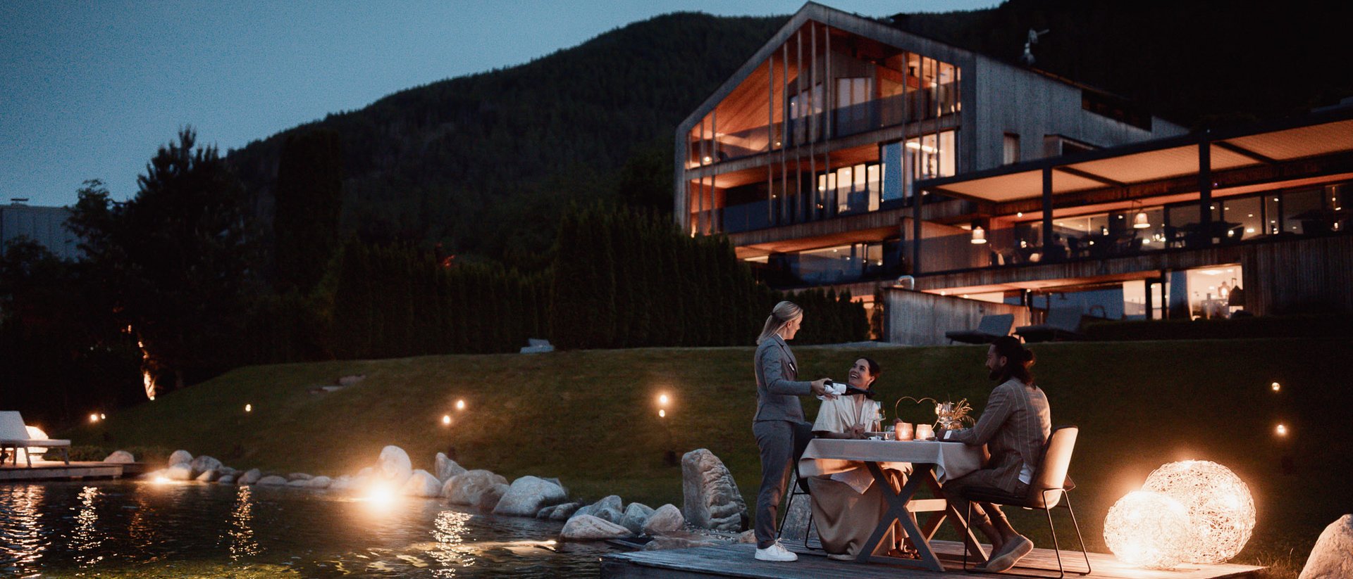 Erfüllender Urlaub in Ihrem Luxus-Chalet in Südtirol Abendessen am See vor modernem Haus in den Bergen bei Dämmerung