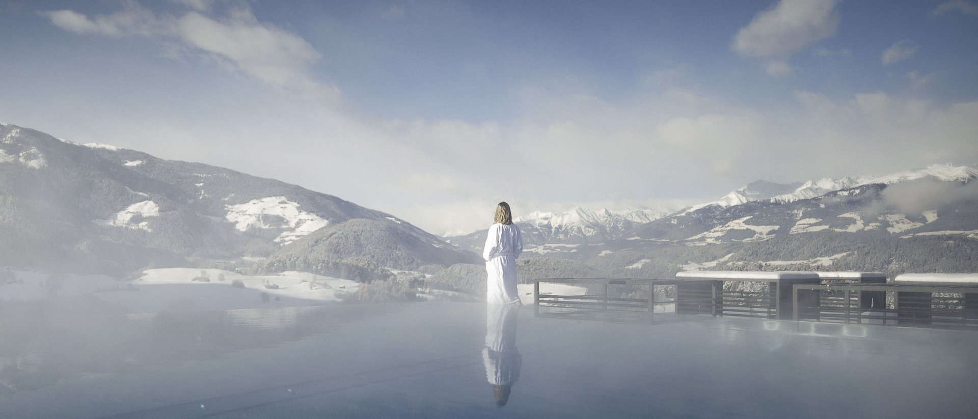 Die Quelle der Erfüllung: Ihr Hotel am Kronplatz Person in Bademantel blickt auf schneebedeckte Berge von einem Pool aus