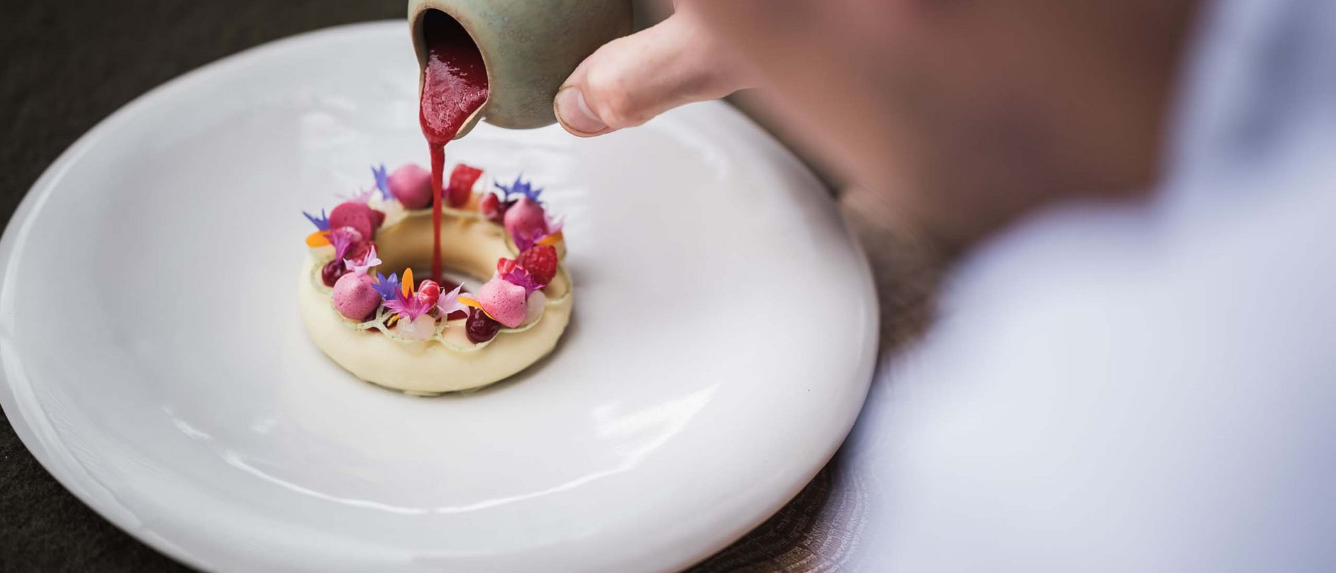 Erfüllender Urlaub in Ihrem Luxus-Chalet in Südtirol Hand gießt rote Sauce auf kunstvoll dekoriertes Dessert auf weißem Teller