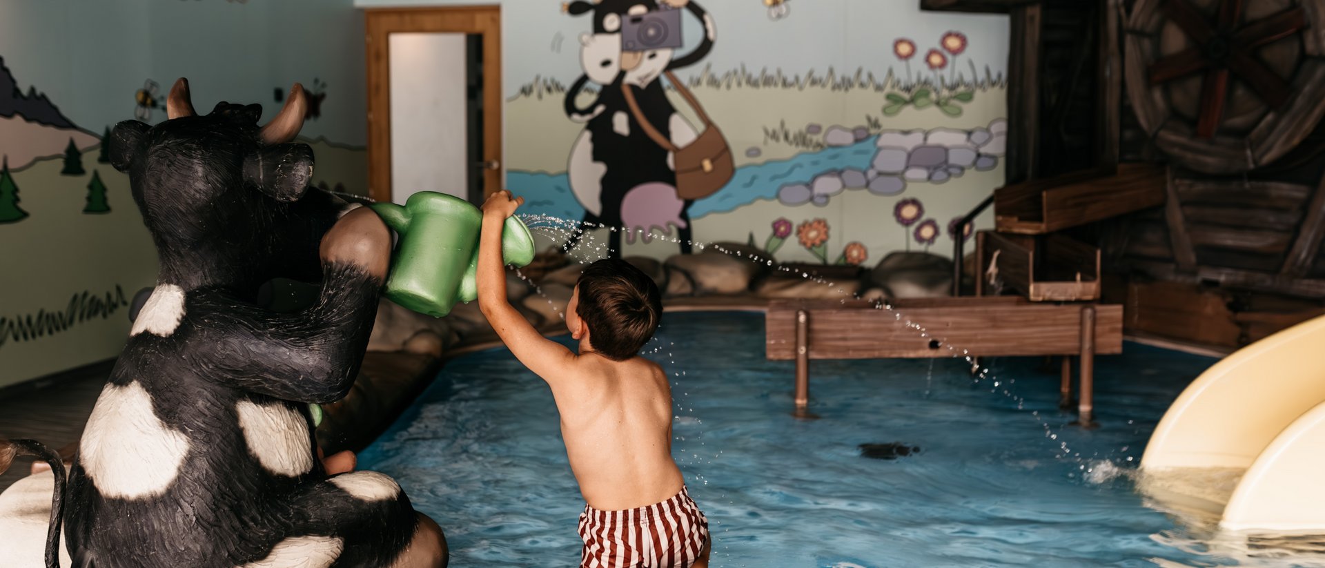 Die Quelle der Erfüllung: Ihr Hotel am Kronplatz Kind spielt mit Gießkanne neben Kuhfigur in einem Innenpool mit Wandmalerei