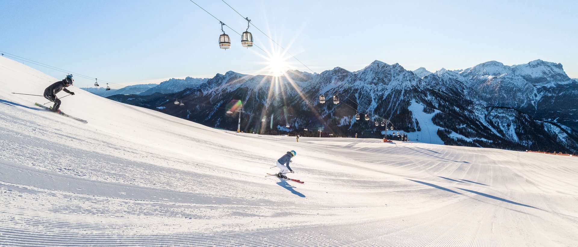 Die Quelle der Erfüllung: Ihr Hotel am Kronplatz Zwei Skifahrer auf präparierter Piste mit Bergseilbahn und Sonnenuntergang