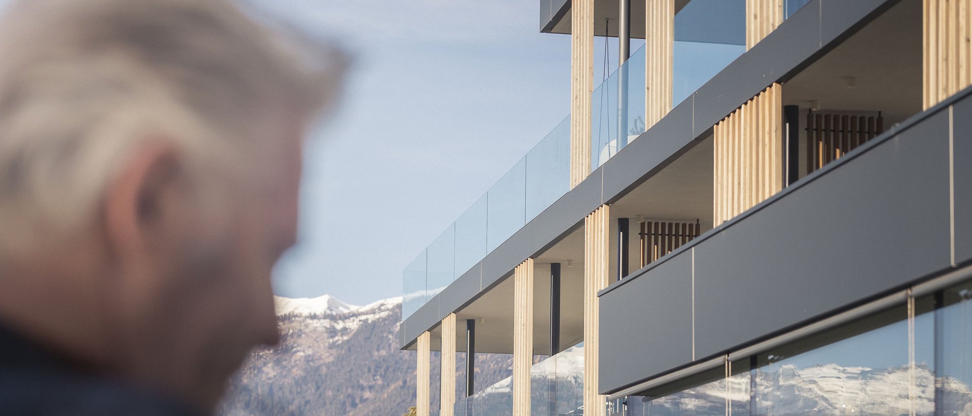 Die Quelle der Erfüllung: Ihr Hotel am Kronplatz Moderner Wohngebäudekomplex mit Bergblick und verschwommenem älteren Mann