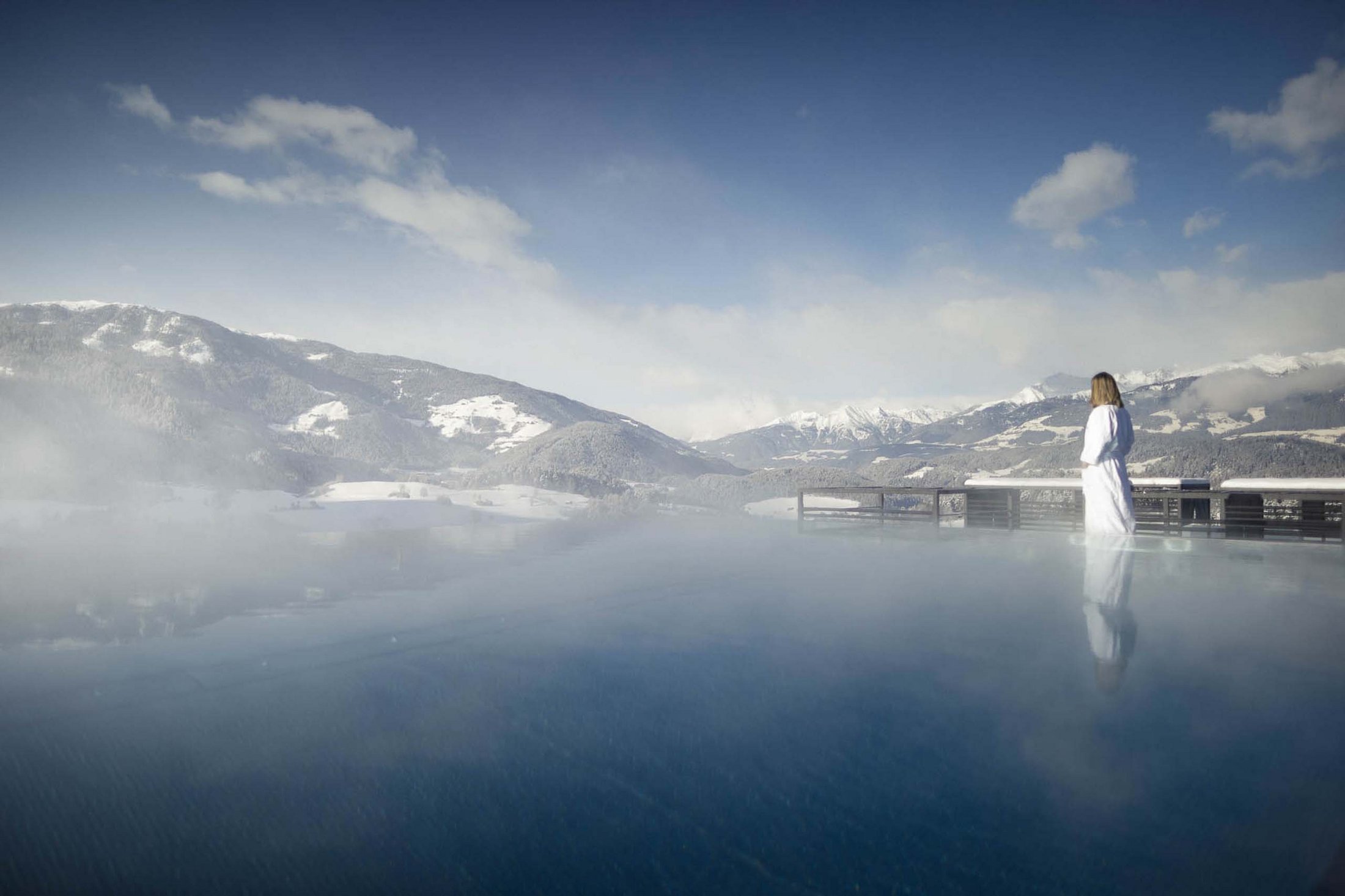 Die Quelle der Erfüllung: Ihr Hotel am Kronplatz Frau in Bademantel am beheizten Außenpool mit Blick auf schneebedeckte Berge