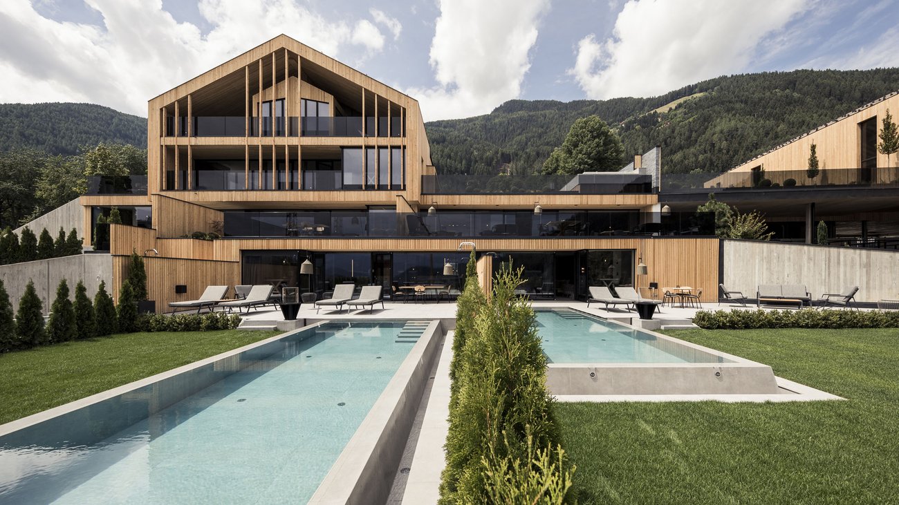 Ihre Hotels in Südtirol: die Heimat der Fülle Modernes Holzhaus mit zwei Pools und Liegestühlen vor Bergen unter bewölktem Himmel