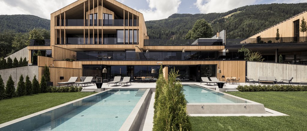 Ihre Hotels in Südtirol: die Heimat der Fülle Modernes Holzhaus mit zwei Pools und Liegestühlen vor Bergen unter bewölktem Himmel