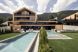 Ihre Hotels in Südtirol: die Heimat der Fülle Modernes Holzhaus mit zwei Pools und Liegestühlen vor Bergen unter bewölktem Himmel