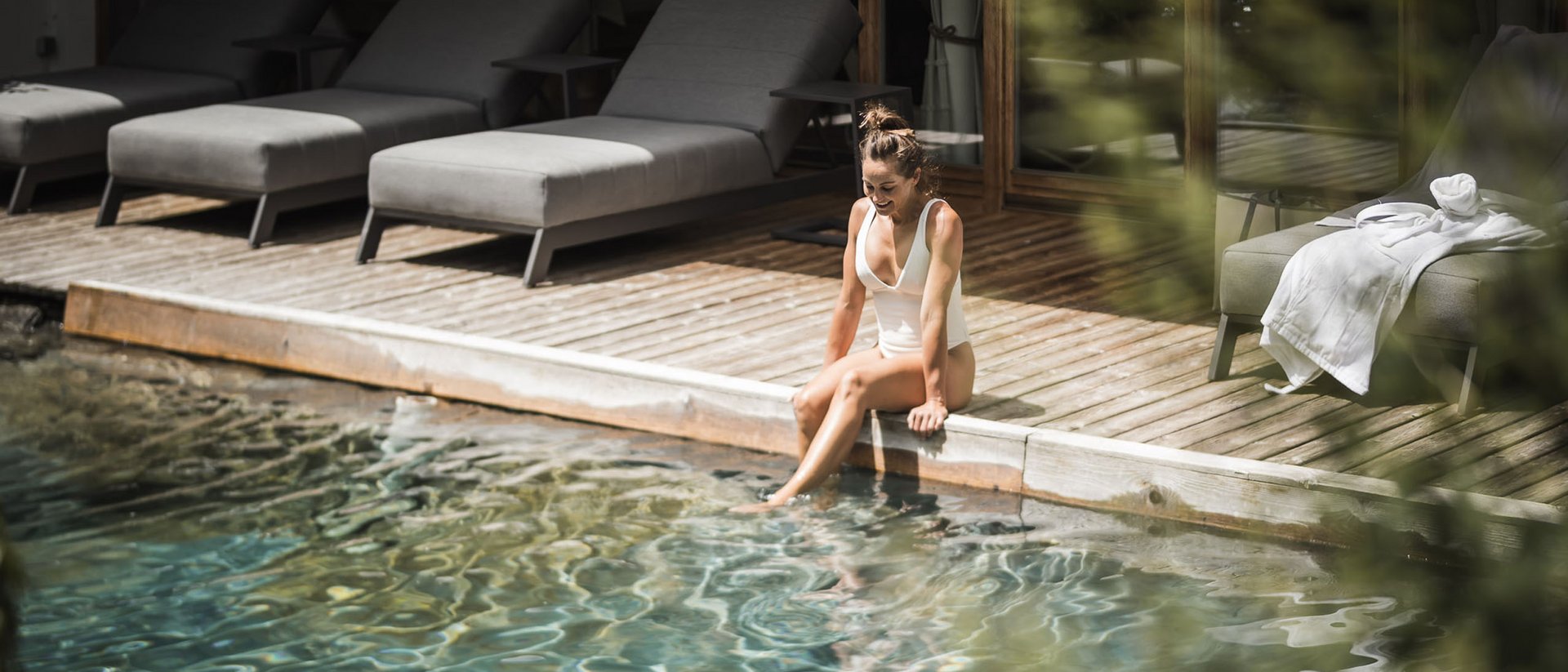 Der Ursprung des Wohlgefühls: Ihr Hotel im Pustertal Frau im weißen Badeanzug sitzt am Pool und taucht ihre Füße ins Wasser
