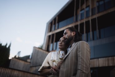 Wir sind Ihre Golfhotels in Südtirol. Paar mit Wein vor modernem Gebäude bei Sonnenuntergang