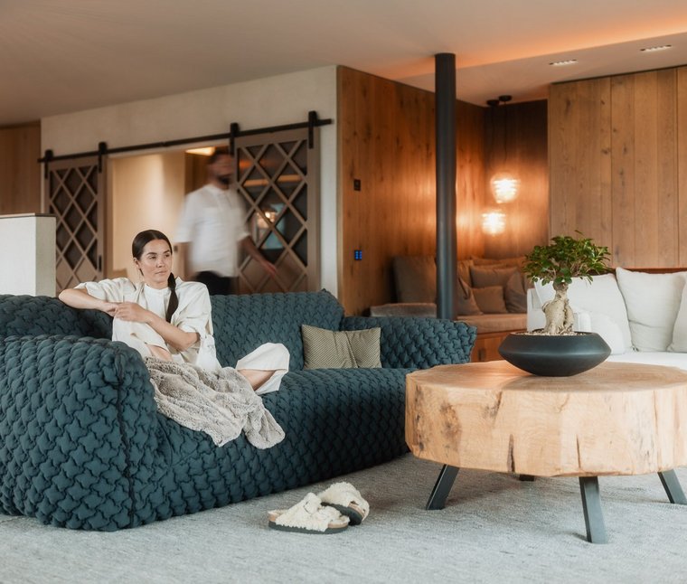 Ihre Hotels in Südtirol: die Heimat der Fülle Frau sitzt auf blauem Sofa in modernem Wohnzimmer mit Holztisch und Kamin