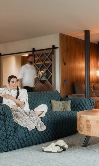 Ihre Hotels in Südtirol: die Heimat der Fülle Frau sitzt auf blauem Sofa in modernem Wohnzimmer mit Holztisch und Kamin