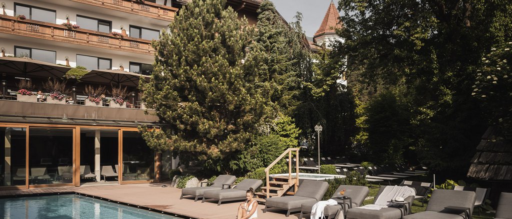 Ihre Hotels in Südtirol: die Heimat der Fülle Frau sitzt am Pool vor Hotel mit Liegestühlen und Bäumen