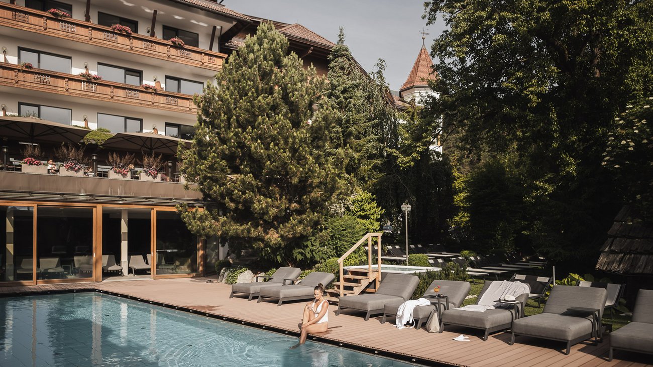 Ihre Hotels in Südtirol: die Heimat der Fülle Frau sitzt am Pool vor Hotel mit Liegestühlen und Bäumen