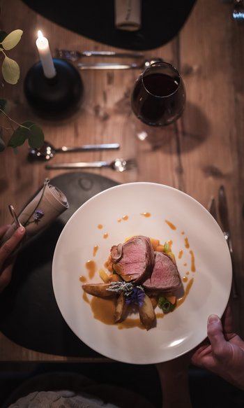 Ihre Hotels in Südtirol: die Heimat der Fülle Gedeckter Tisch mit Kerze, Wein und angerichtetem Fleischgericht auf weißem Teller