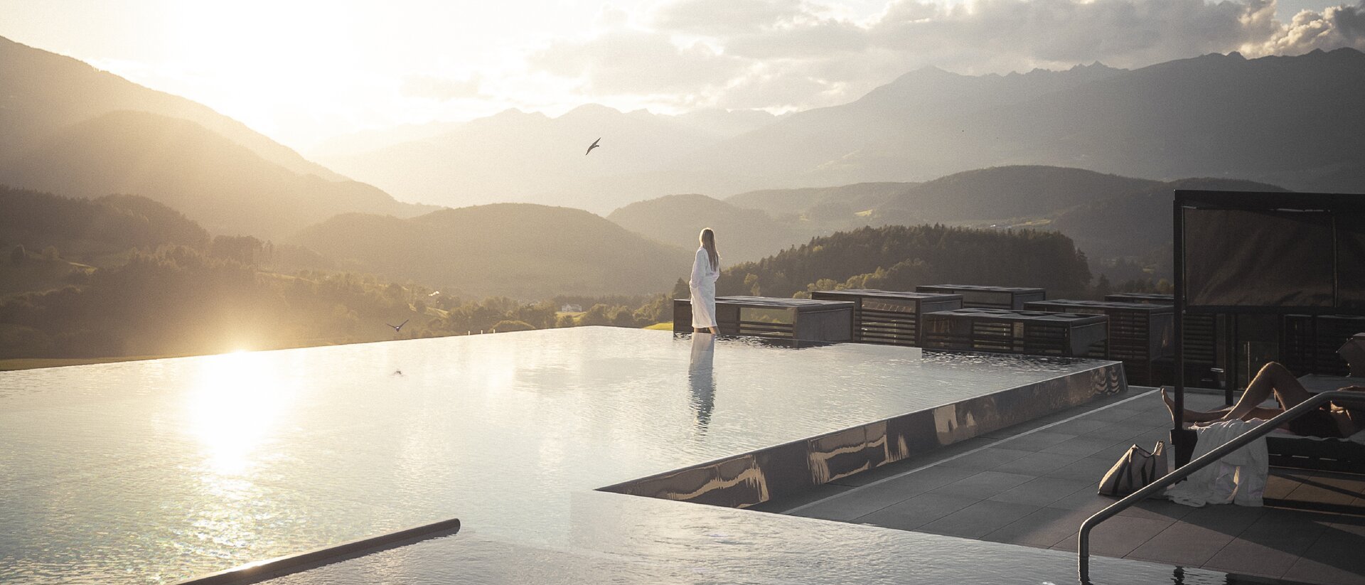 Die Quelle der Erfüllung: Ihr Hotel am Kronplatz Frau in weißem Bademantel am Infinity-Pool mit Bergblick bei Sonnenuntergang