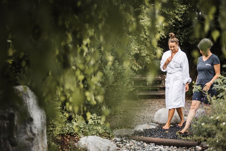 Der Ursprung des Wohlgefühls: Ihr Hotel im Pustertal Zwei Frauen gehen barfuß auf einem Gartenweg mit Steinen umgeben von Pflanzen