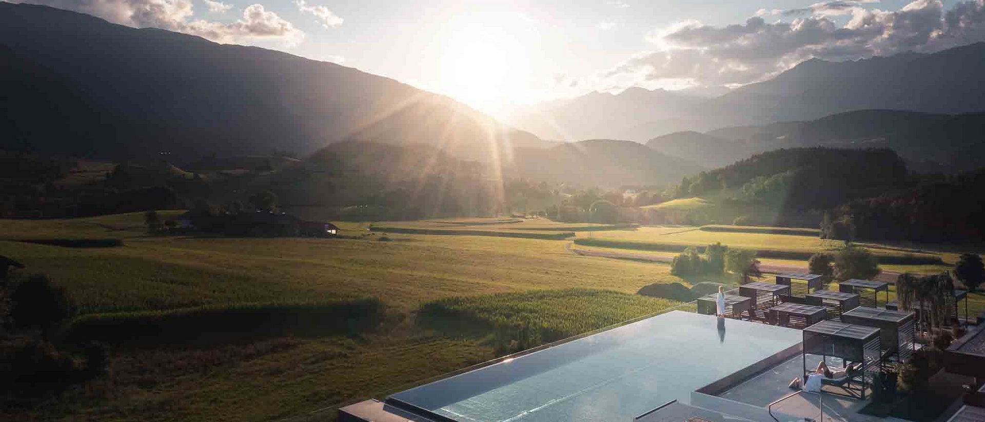 Die Quelle der Erfüllung: Ihr Hotel am Kronplatz Sonnenuntergang über Bergtal mit Feldern und Infinity-Pool