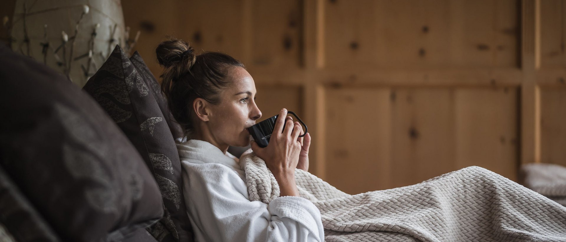 Der Ursprung des Wohlgefühls: Ihr Hotel im Pustertal Frau in Bademantel trinkt Kaffee auf gemütlichem Sofa mit Decke