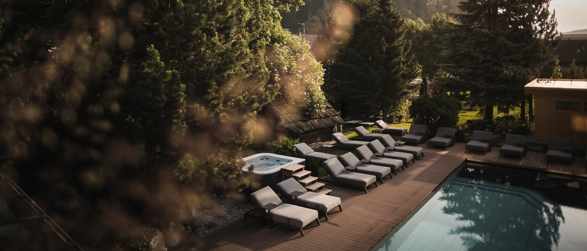 Der Ursprung des Wohlgefühls: Ihr Hotel im Pustertal Pool mit Liegestühlen und Whirlpool in einem bewaldeten Bergresort bei Sonnenuntergang