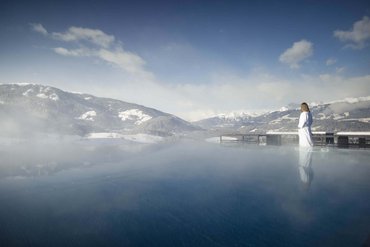 Wir sind Ihre Golfhotels in Südtirol. Frau in Bademantel am beheizten Außenpool mit Blick auf schneebedeckte Berge
